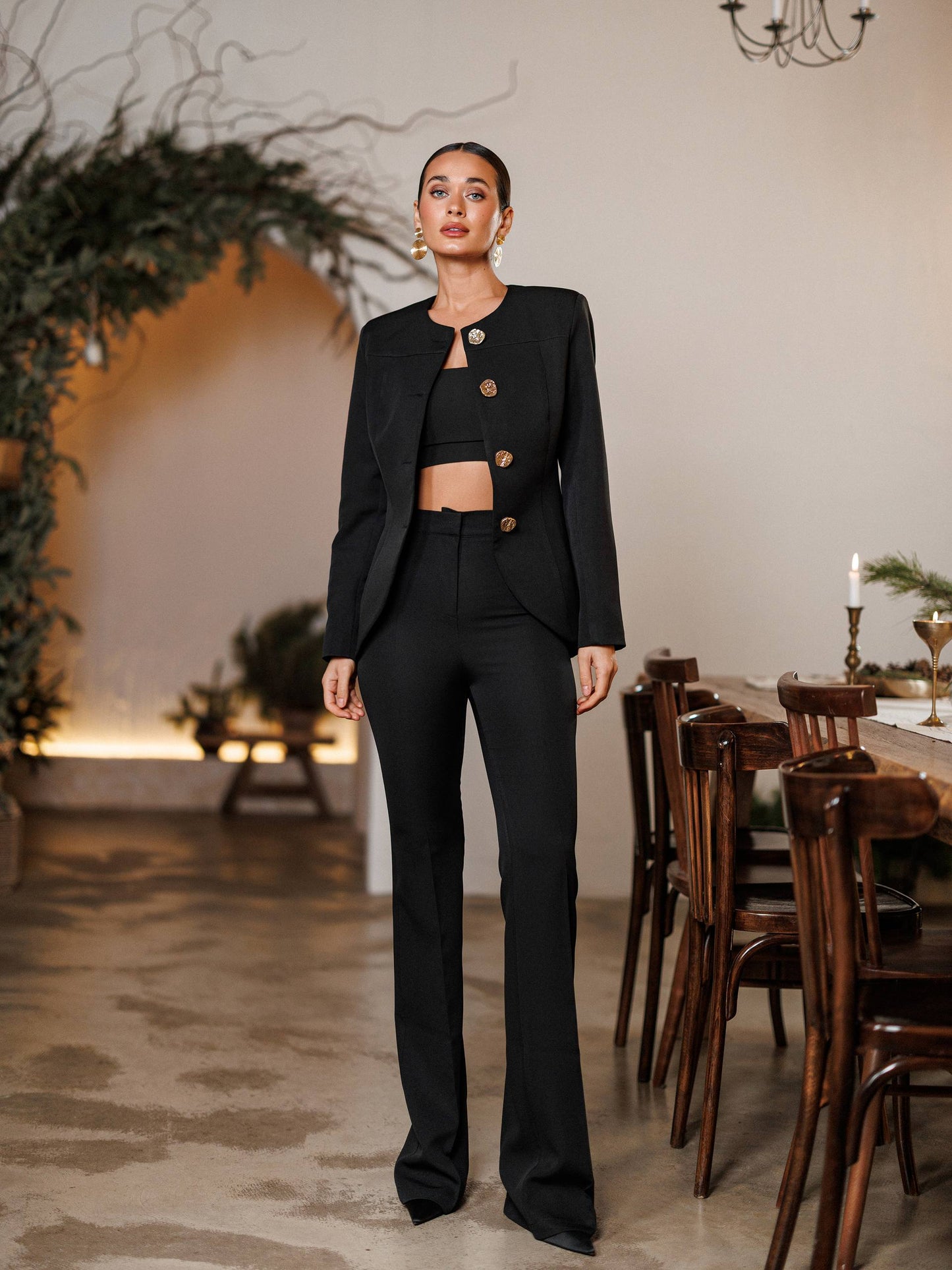 Woman in a black suit standing in a room with wooden furniture and plants.