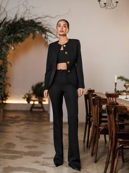 Woman in a black suit standing in a room with wooden furniture and plants.
