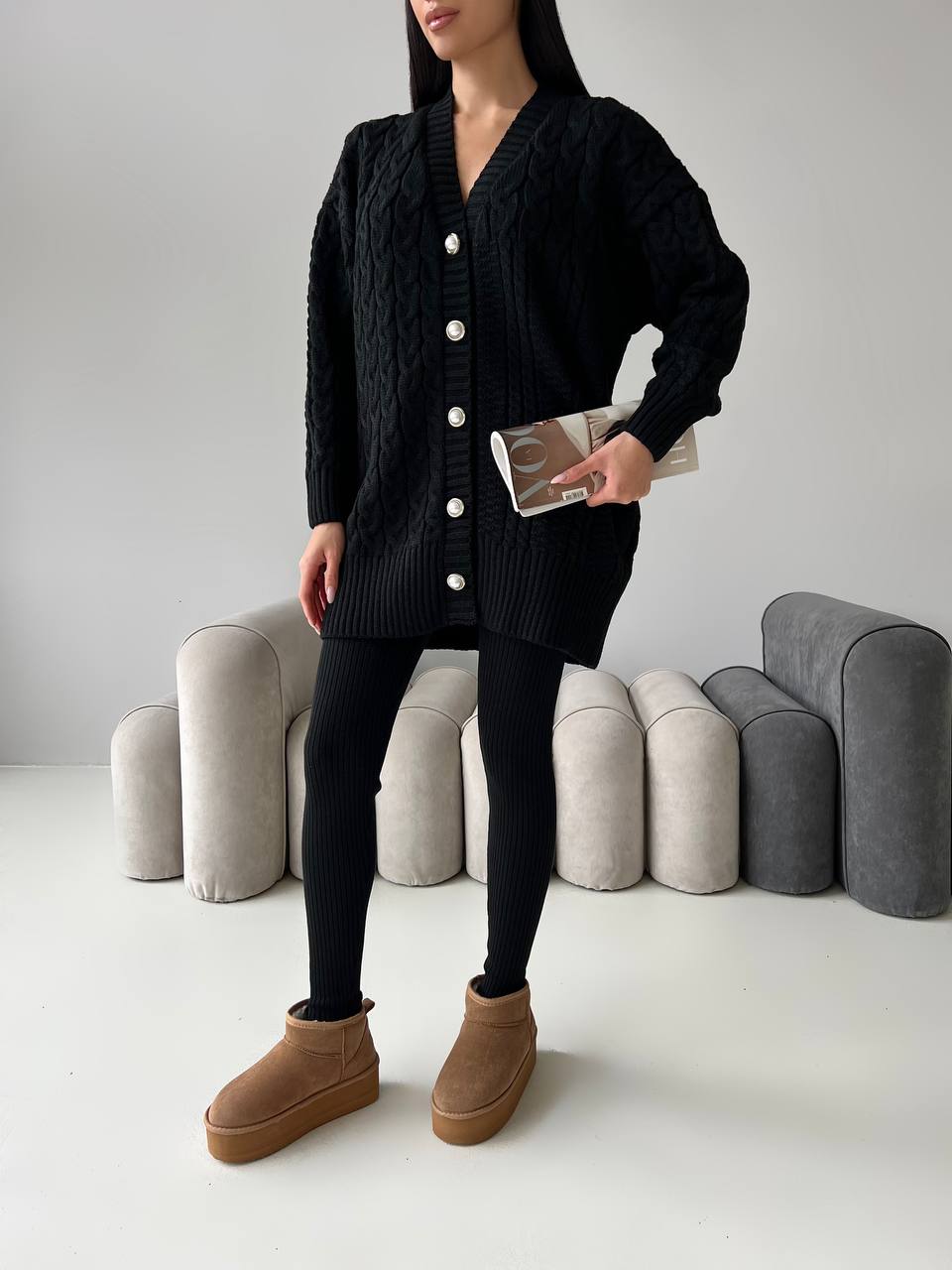 Person wearing a black cardigan, black leggings, and brown boots against a neutral background