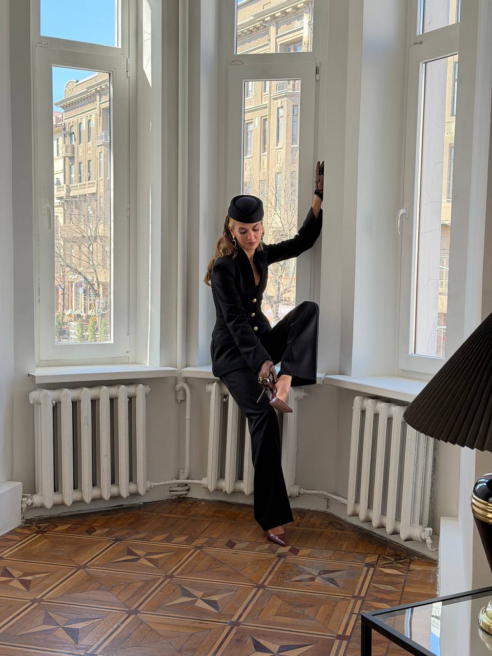 Person in a black outfit sitting on a window sill in a room with large windows and wooden floor.