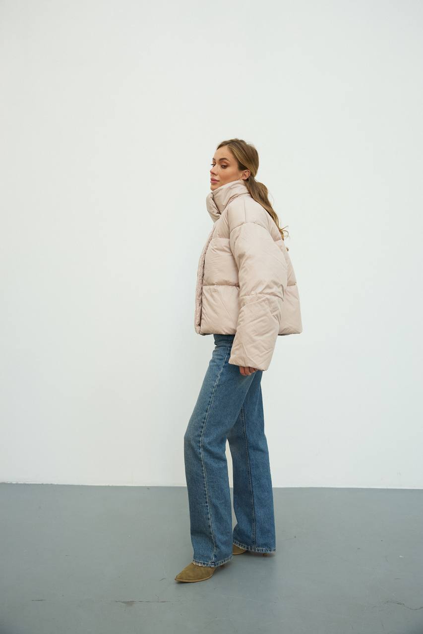 Person wearing a beige puffer jacket and blue jeans standing against a white wall.