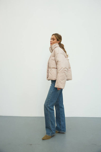 Person wearing a beige puffer jacket and blue jeans standing against a white wall.