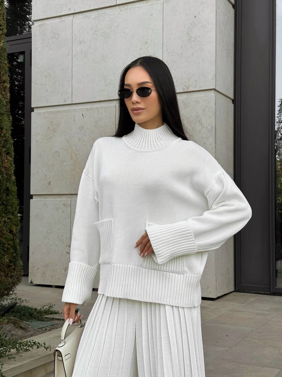 Woman wearing a white sweater and skirt standing against a light-colored wall.