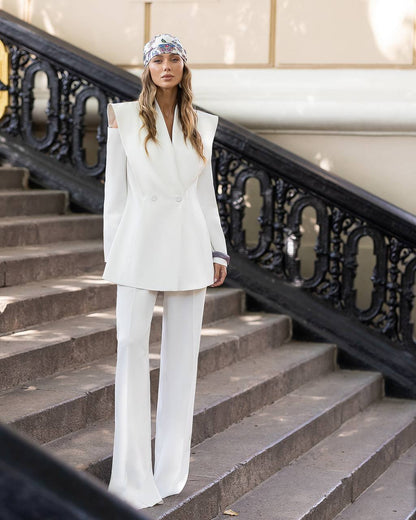 A woman stands on outdoor stone stairs wearing a white sleeveless blazer and matching wide-leg trousers, with a patterned headscarf. A black ornate railing and beige building wall are visible behind her.