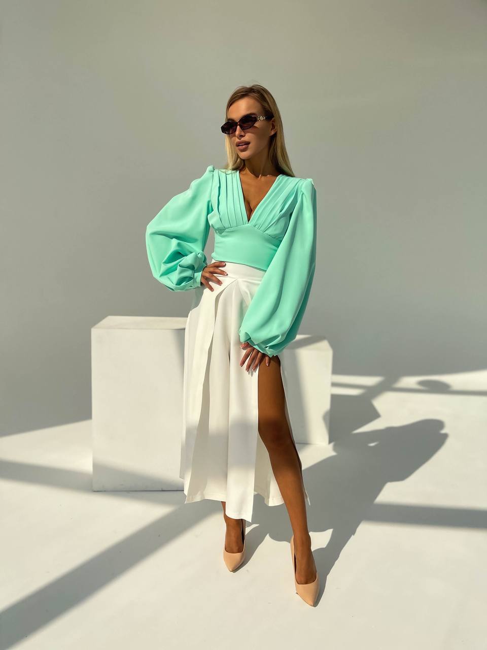 Woman wearing a light green blouse with puffed sleeves and a white skirt against a neutral background