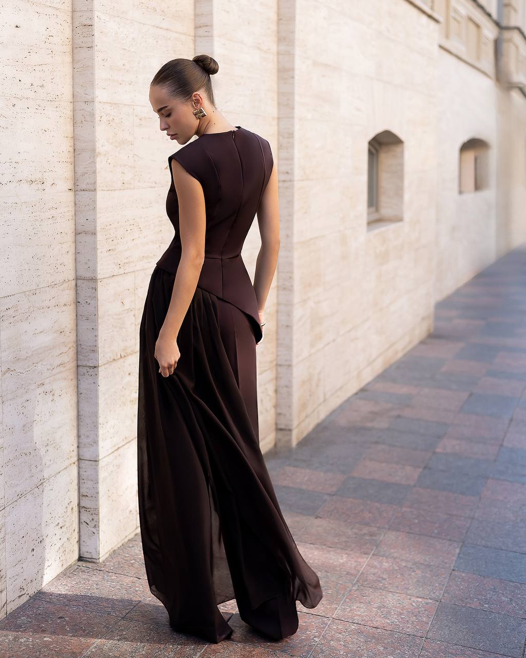 Woman in a dark brown dress standing against a light stone wall.