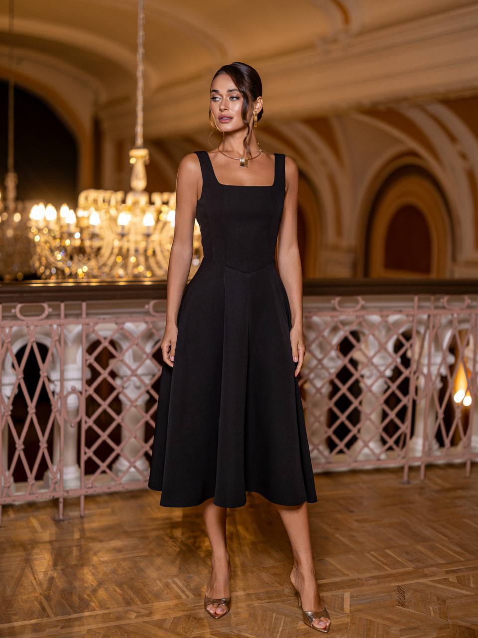 Woman in a black dress standing in an elegant indoor setting with chandeliers.
