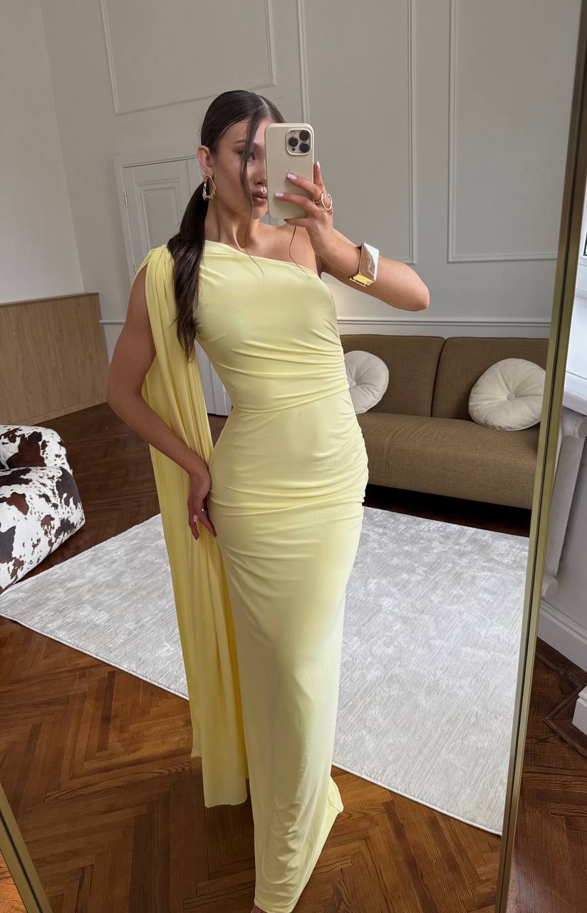 Woman in a yellow one-shoulder dress taking a mirror selfie in a room with wooden flooring and a couch.