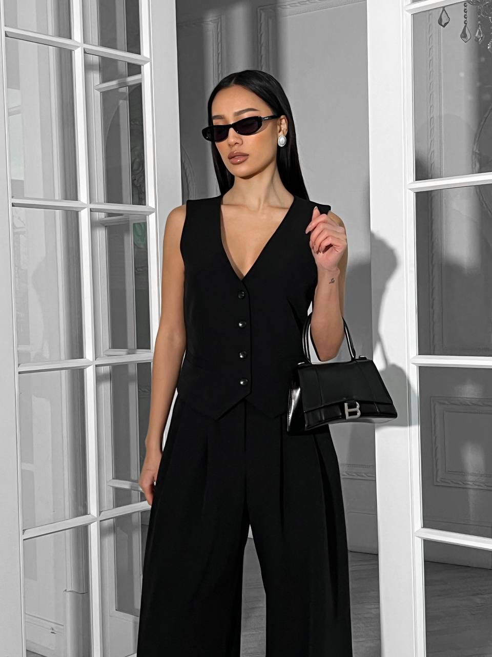 Woman in a black outfit holding a matching handbag in a room with white walls and glass doors.