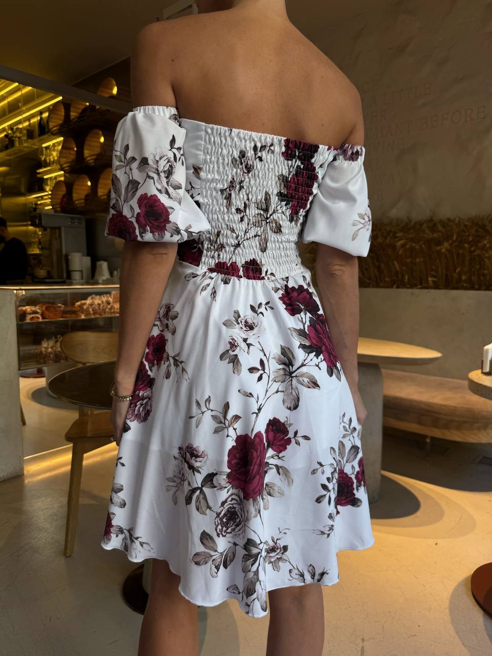 Person wearing a white off-shoulder dress with floral patterns in an indoor setting.