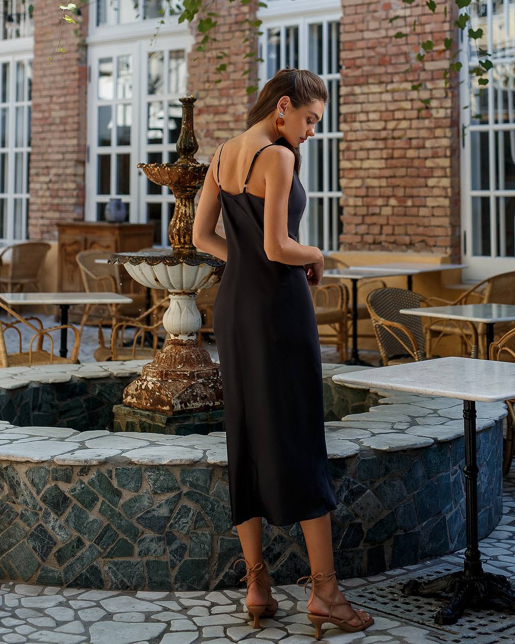 Woman in a black dress standing near a fountain in an outdoor setting with tables and chairs.
