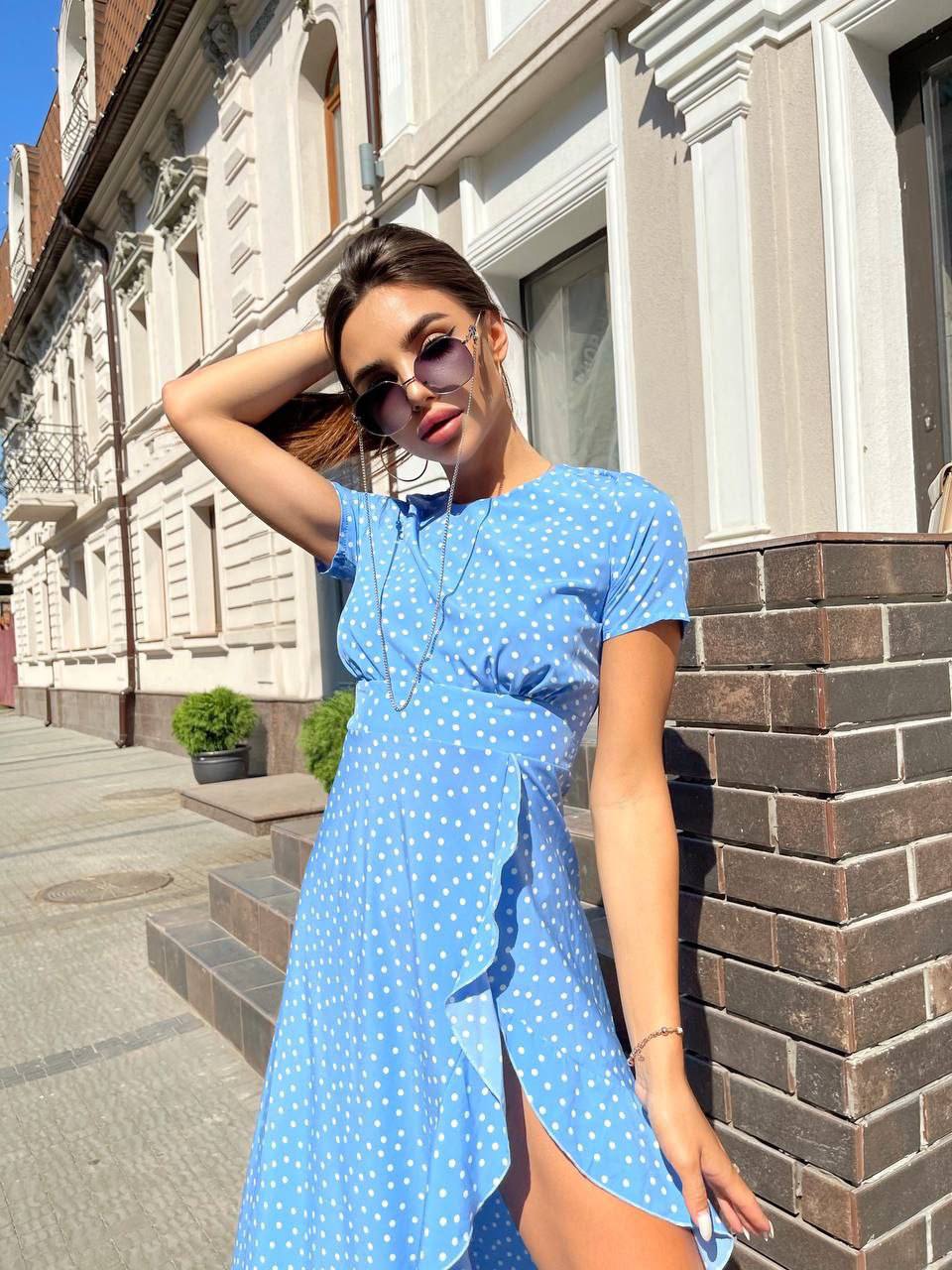 Woman in a blue polka dot dress standing against a brick wall.