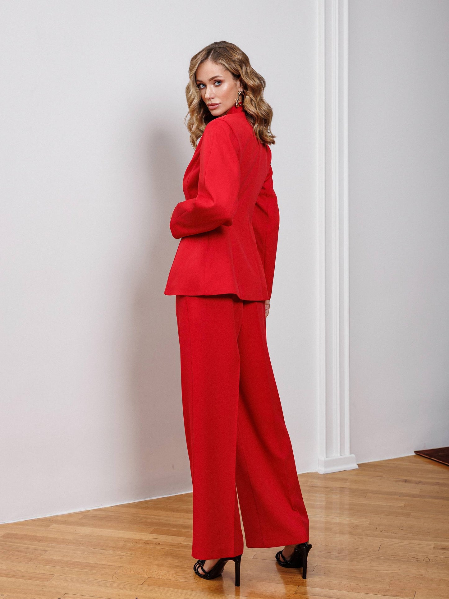 Woman wearing a red suit standing in a room with wooden floor and white walls.