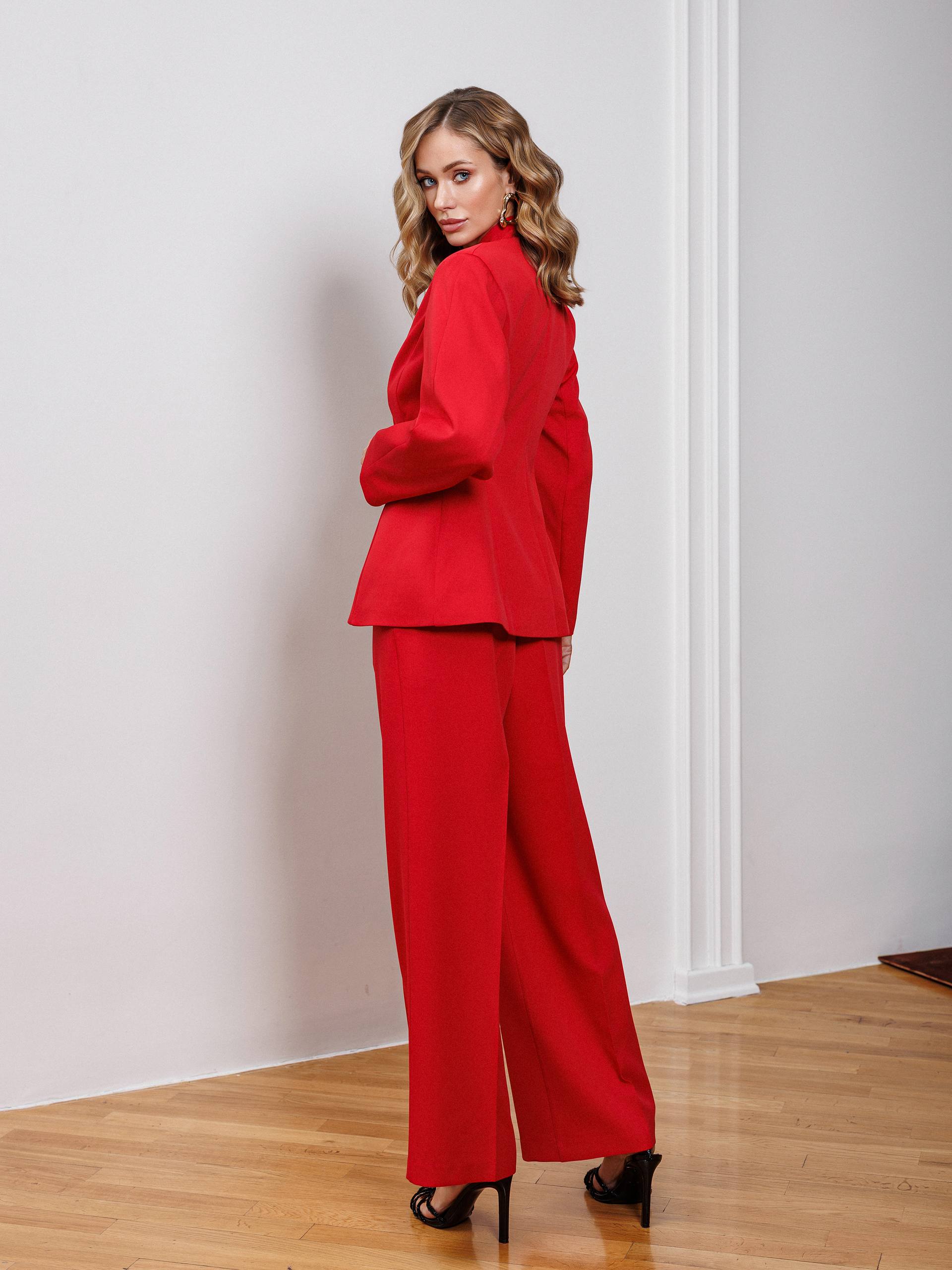 Woman wearing a red suit standing in a room with wooden floor and white walls.