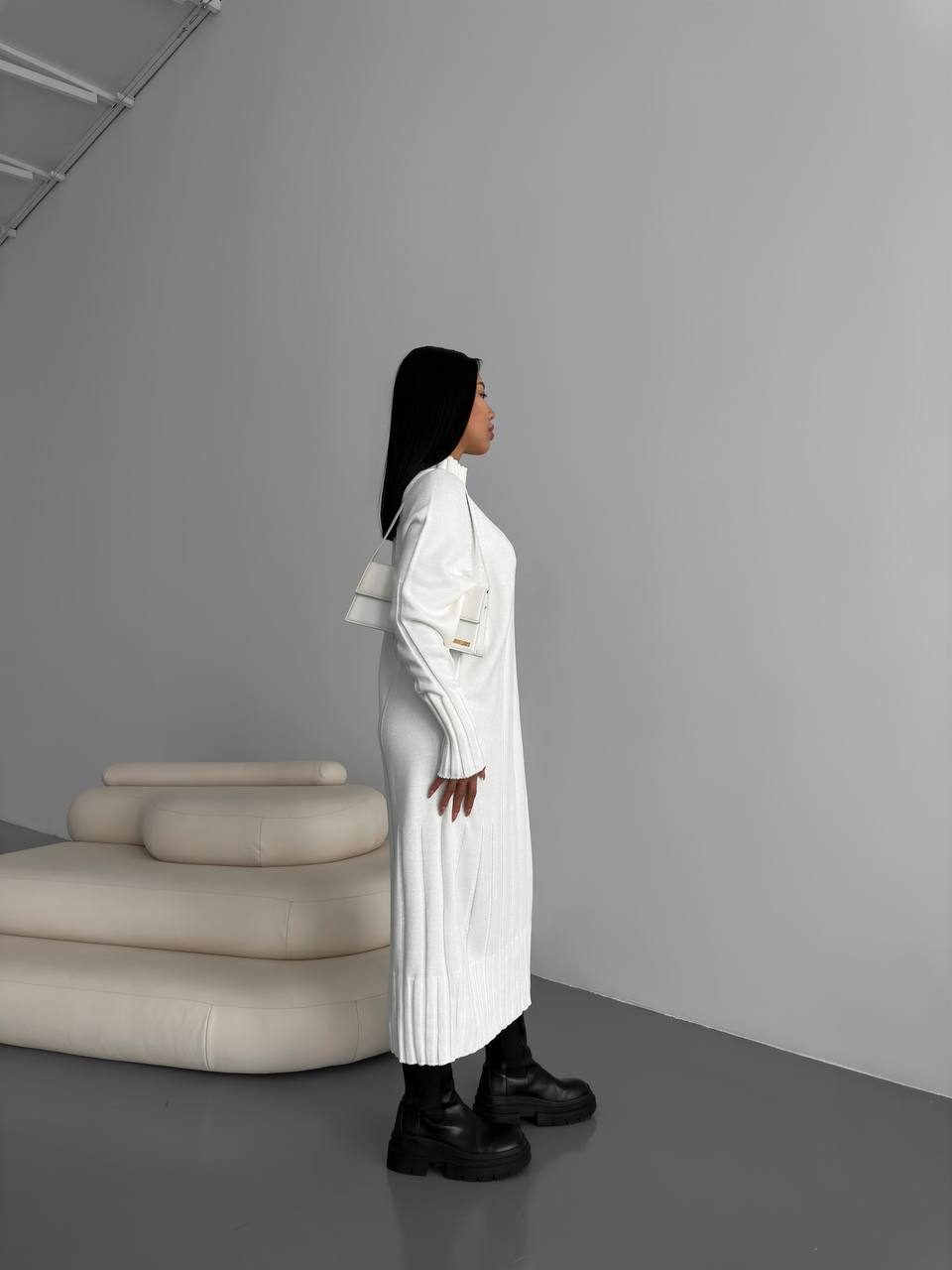 Person wearing a long white coat standing in a minimalistic room with gray walls and a beige sofa.
