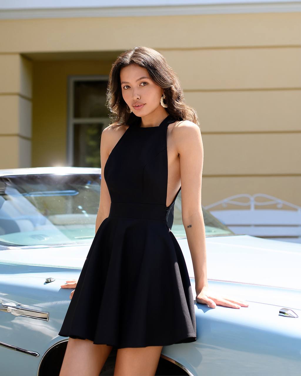 Woman in a black dress leaning on a classic car with a yellow building in the background