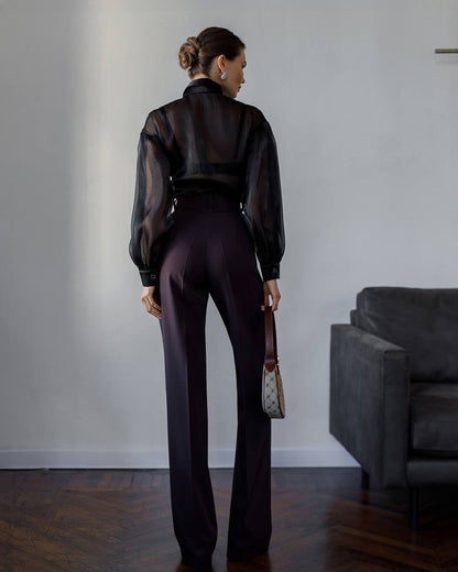 Woman in a black sheer blouse and dark pants standing in a room with a gray sofa.
