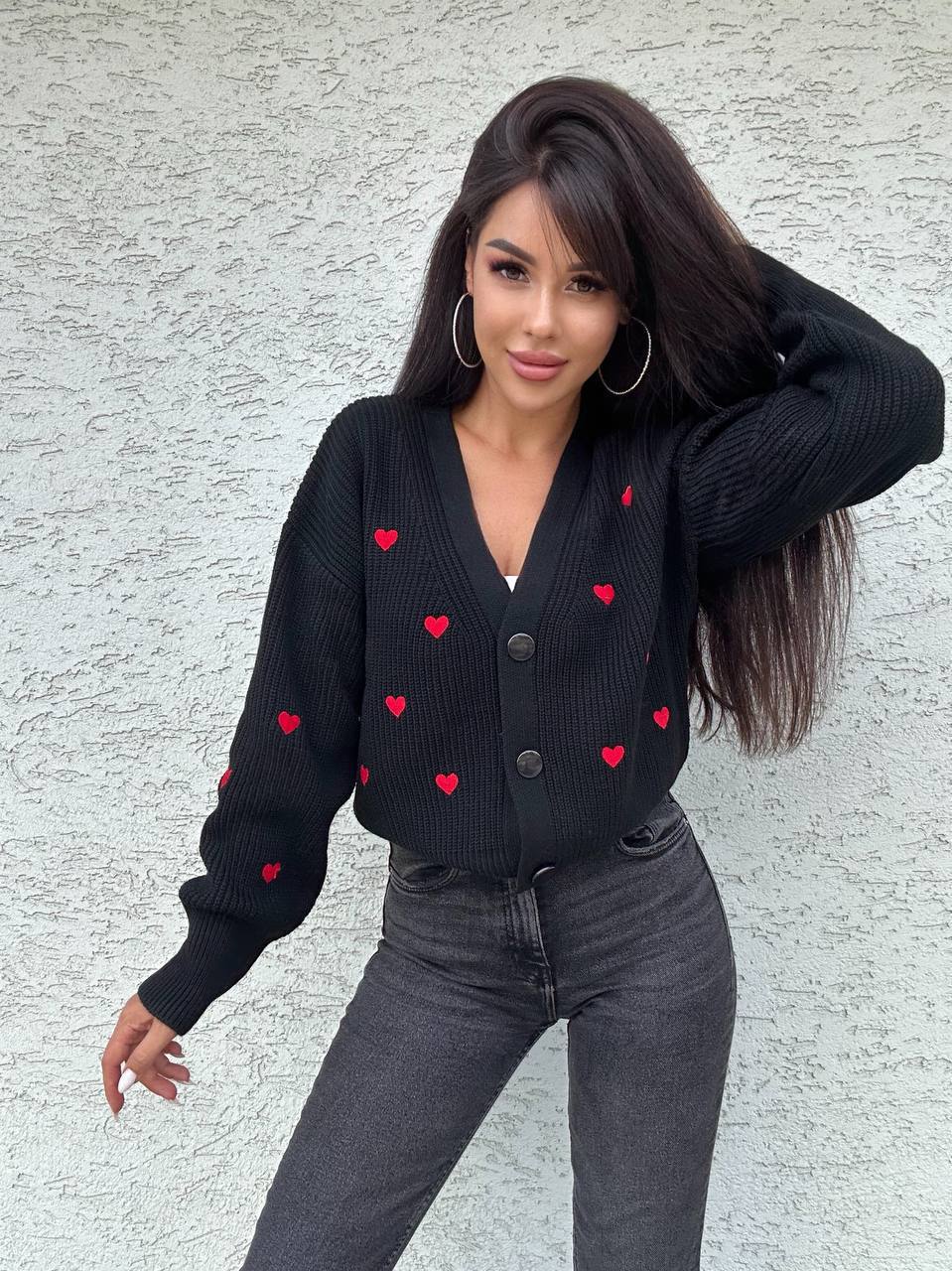 Woman wearing a black cardigan with red heart patterns against a light gray wall.