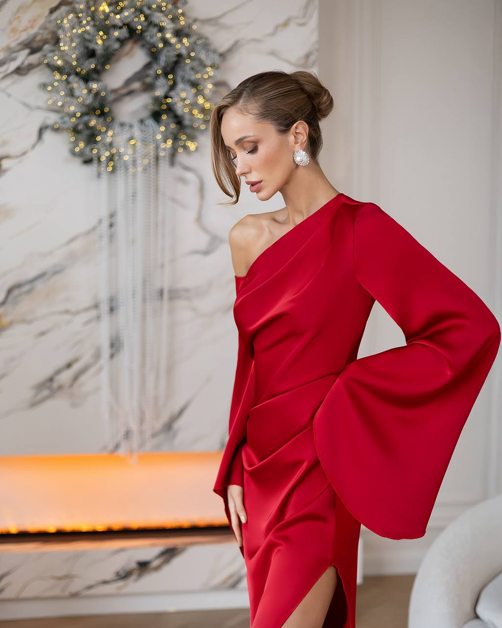 A woman wearing a vibrant red one-shoulder dress with dramatic bell sleeves, posed in front of a marble wall decorated with a holiday wreath.