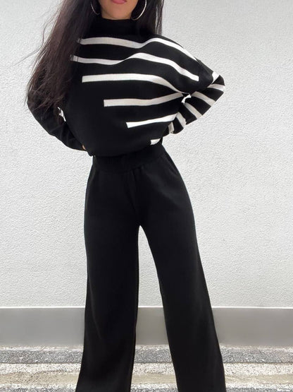 Person wearing a black and white striped sweater and black pants against a light gray wall.