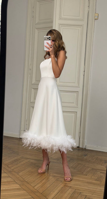 Woman wearing a white dress with feather details in a room with wooden flooring and white walls.