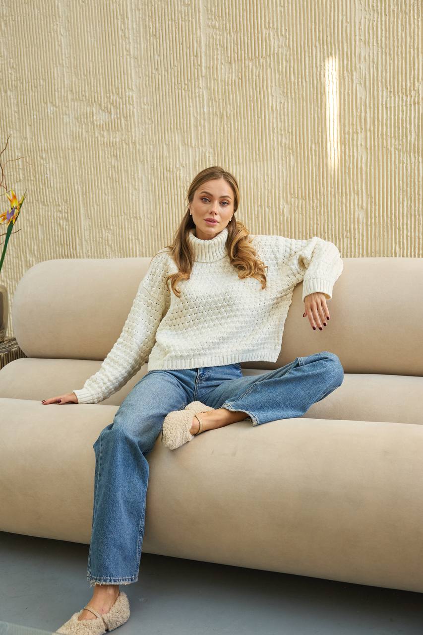 Woman sitting on a beige sofa wearing a white sweater and blue jeans in a room with a textured wall.