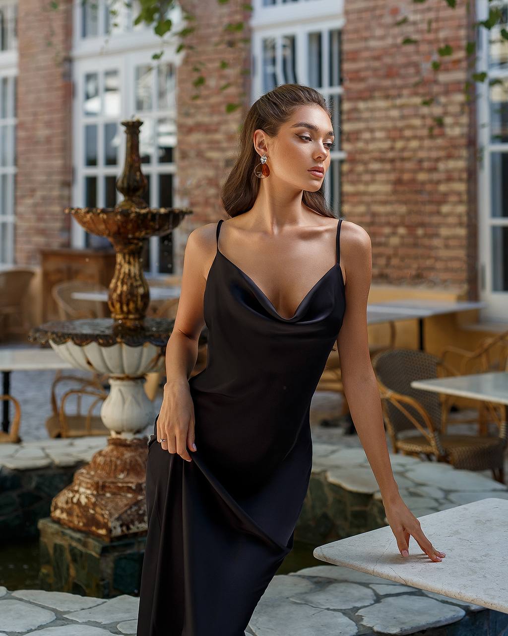 Woman in a black dress standing outdoors near a fountain and tables.