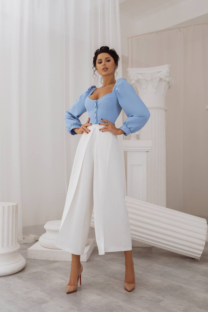 Woman in a light blue top and white pants posing in a stylish interior setting.