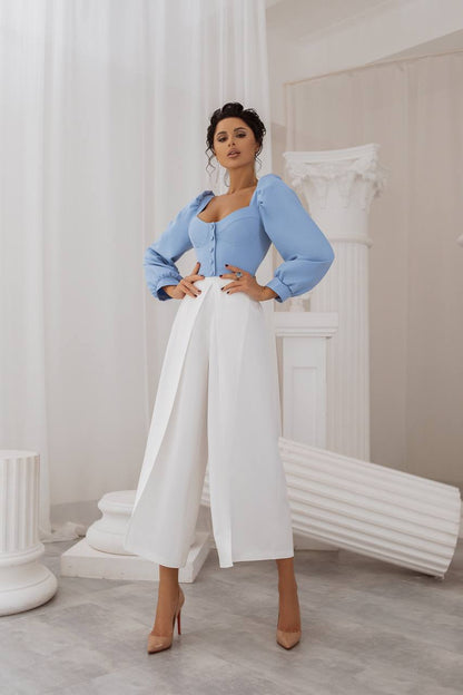 Woman in a light blue top and white pants posing in a stylish interior setting.