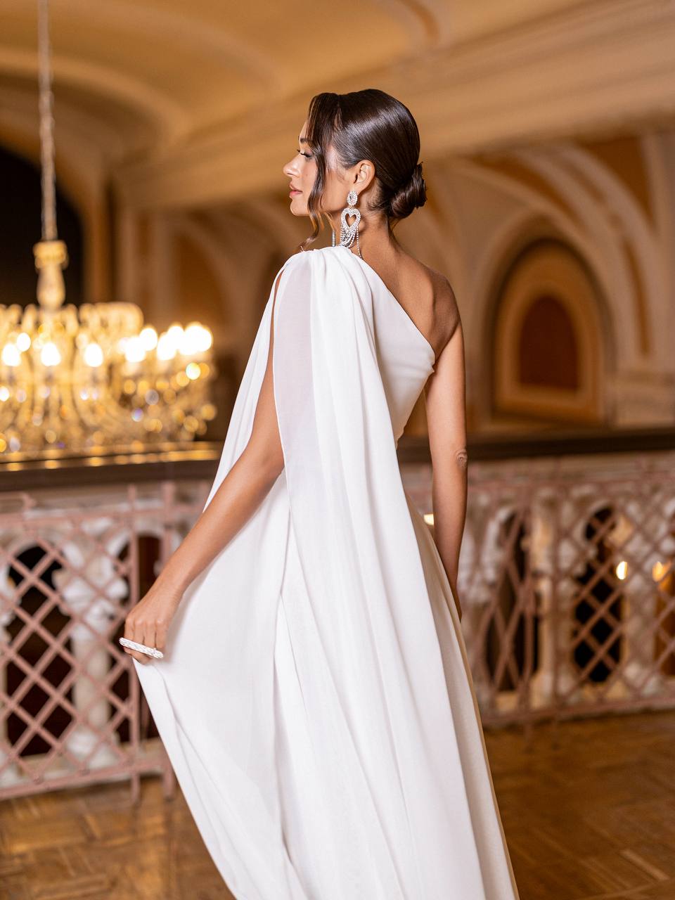 Woman in a white dress standing in an elegant indoor setting with chandeliers.