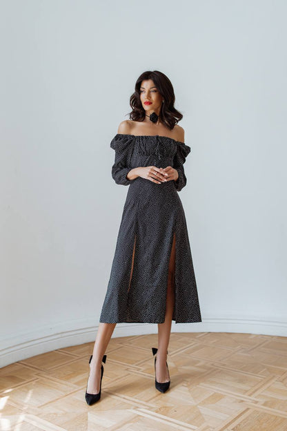 Woman wearing a dark gray off-shoulder dress with a slit, standing in a room with wooden flooring.