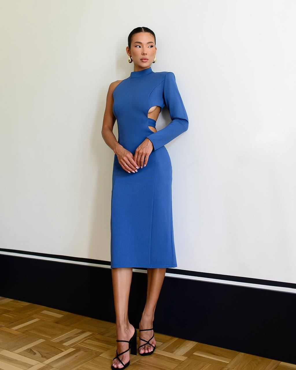 Woman wearing a blue dress with cut-out design against a white wall.