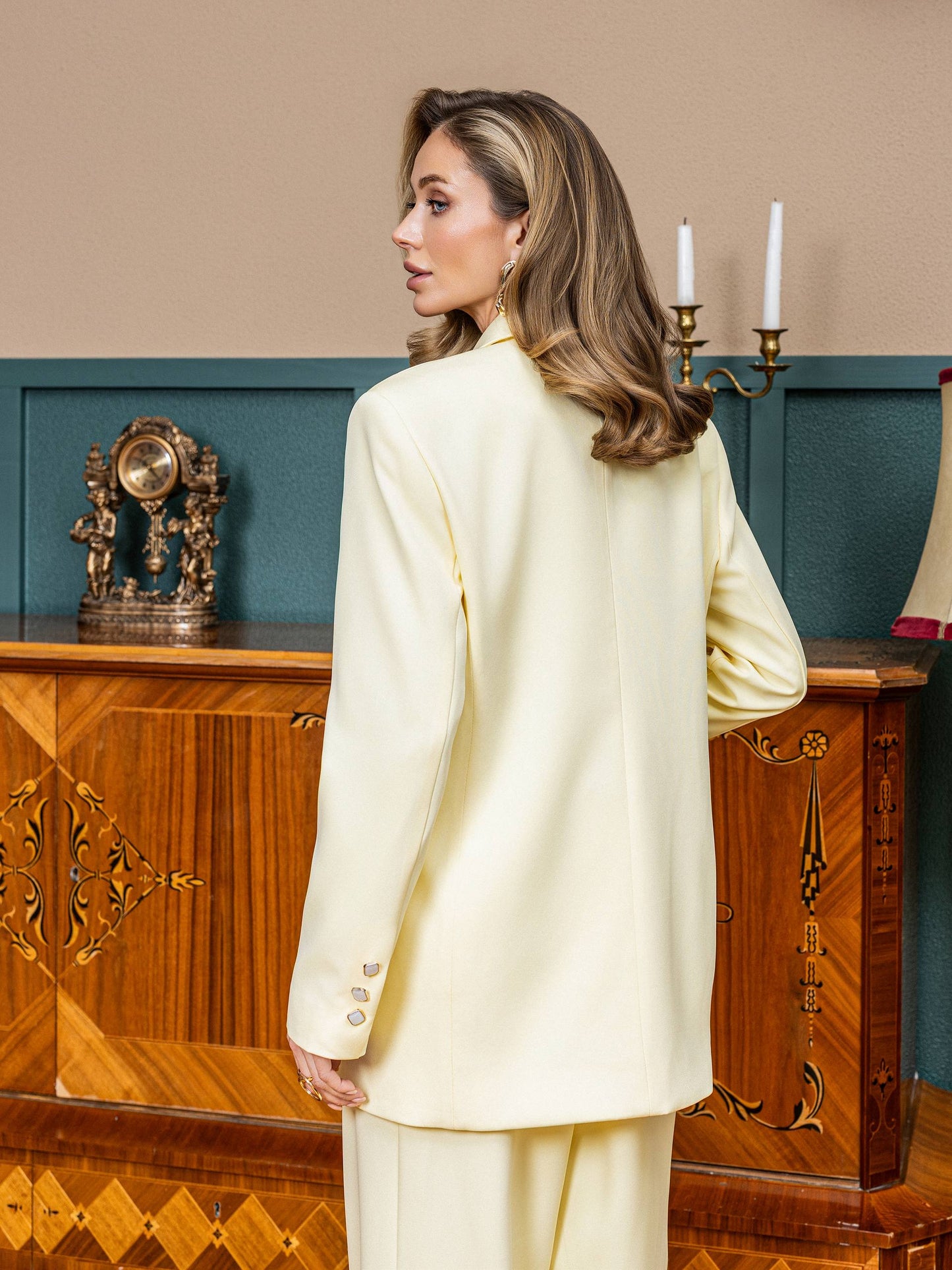Woman in a light-colored suit standing in a room with wooden furniture and decorative items.