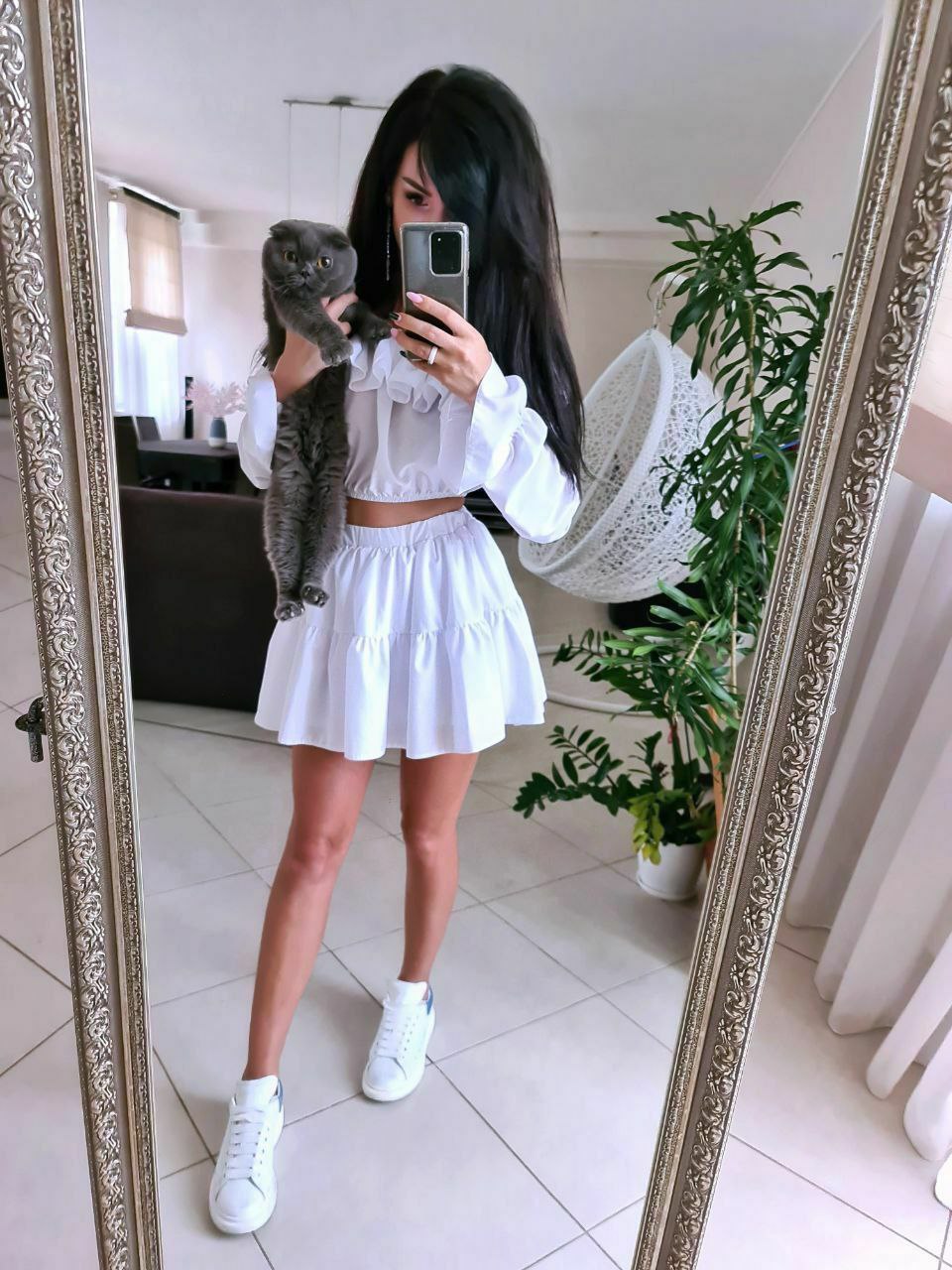 Person taking a mirror selfie wearing a white outfit with a cat on their shoulder, in a room with decor elements.
