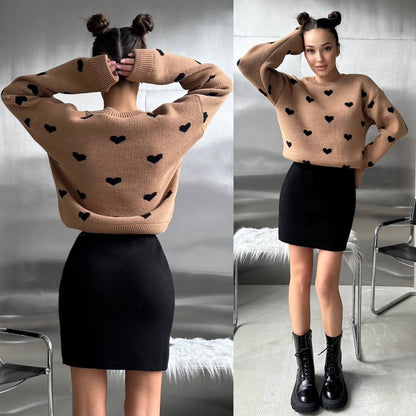 Woman wearing a beige sweater with black heart patterns and a black skirt indoors.