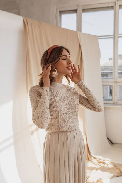 Woman wearing a beige crochet top and pleated skirt in a softly lit room.