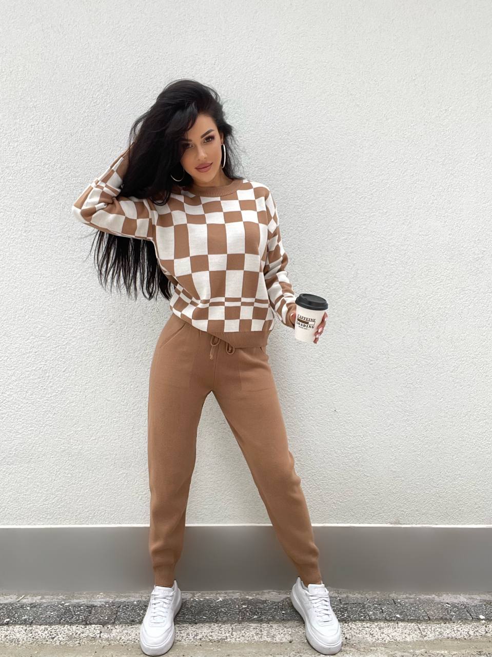 Woman wearing a checkered sweater and brown pants, holding a coffee cup against a plain background