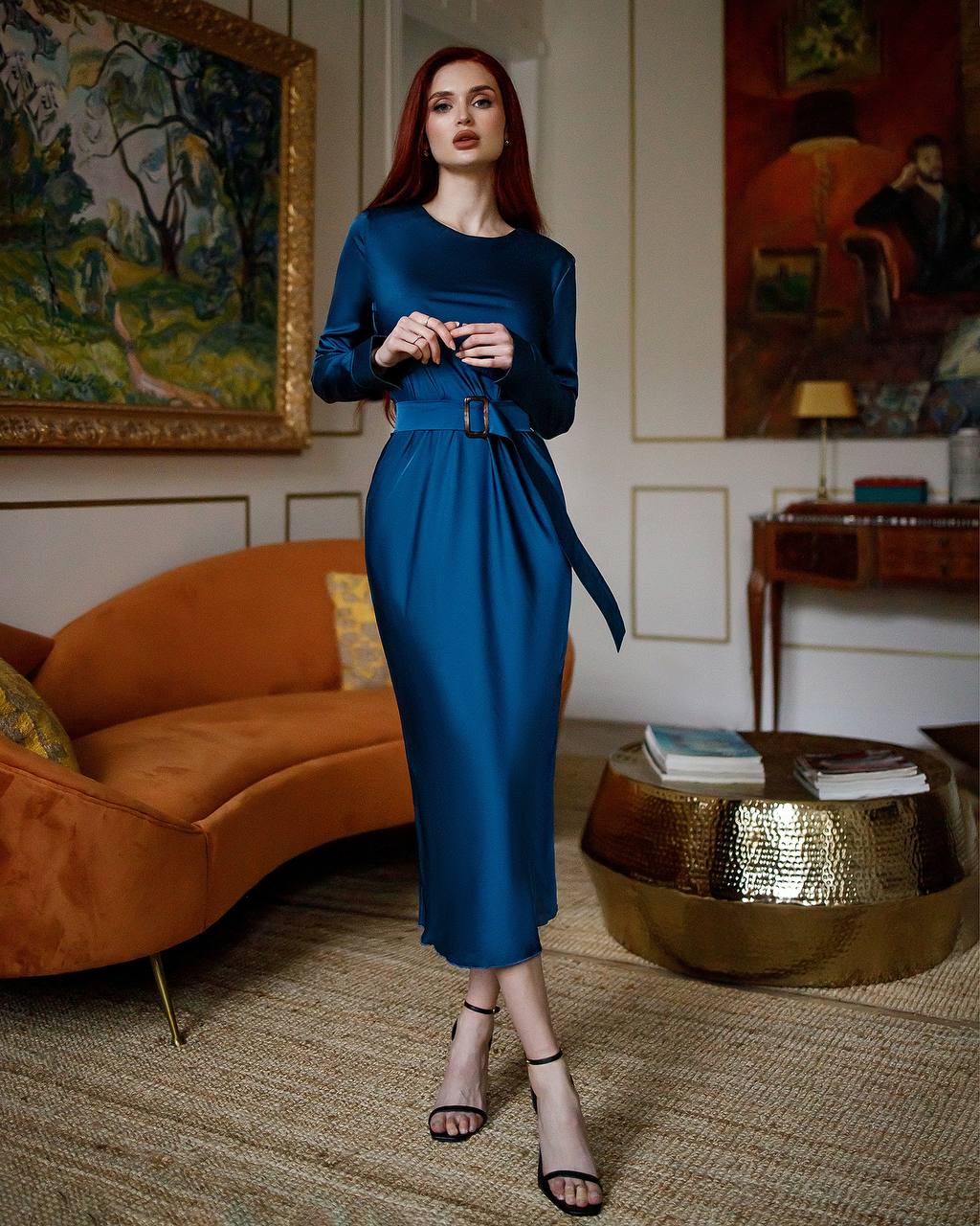 Woman in a blue dress standing in an elegant room with a couch and paintings.