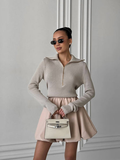 Woman in a beige sweater and skirt holding a white handbag against a gray wall.
