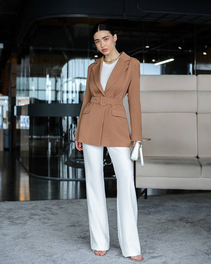 Woman in a brown blazer and white pants standing in an indoor setting.