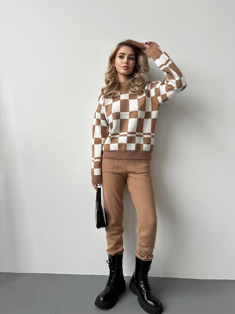 Woman wearing a checkered sweater and tan pants with black boots against a plain background