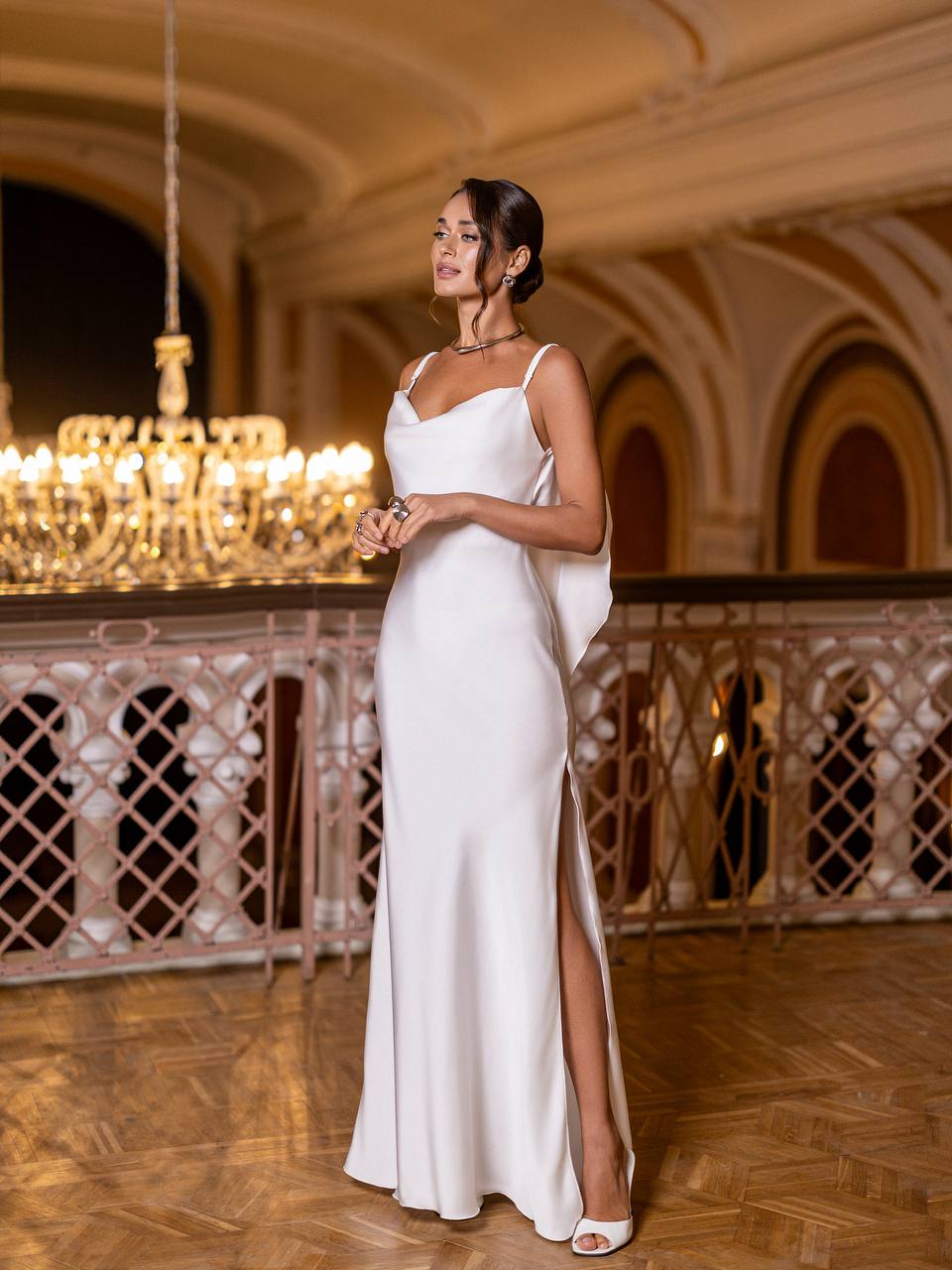 Woman in a white evening gown standing in an elegant indoor setting with chandeliers.