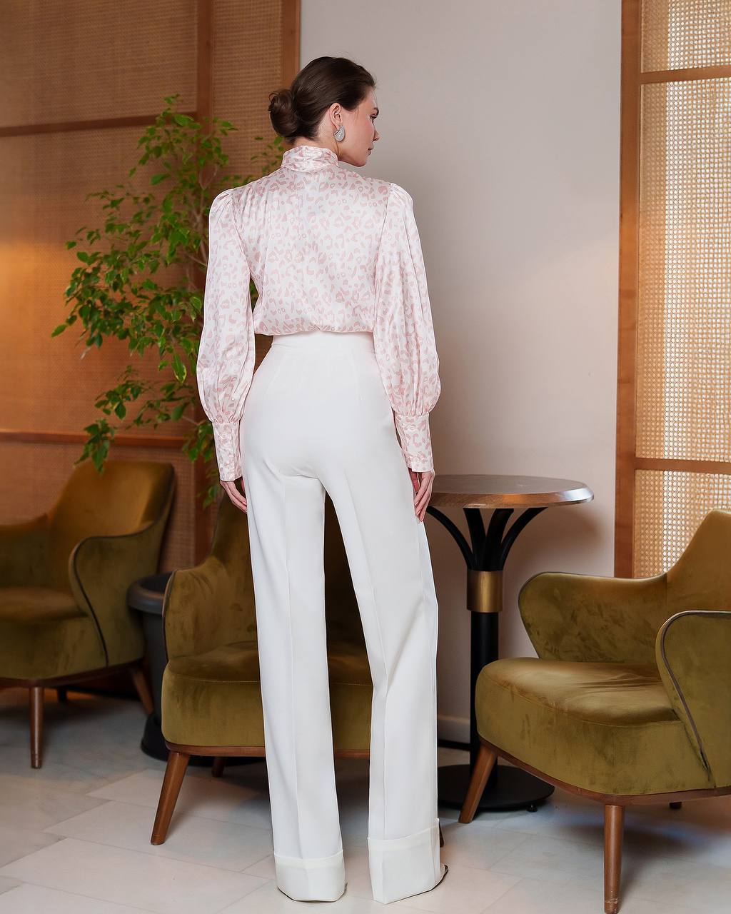 Woman in a light blouse and white pants standing in a room with green chairs and a plant.