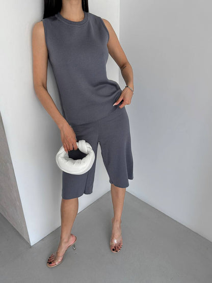 Woman in gray sleeveless top and capri pants holding a white clutch against a plain background