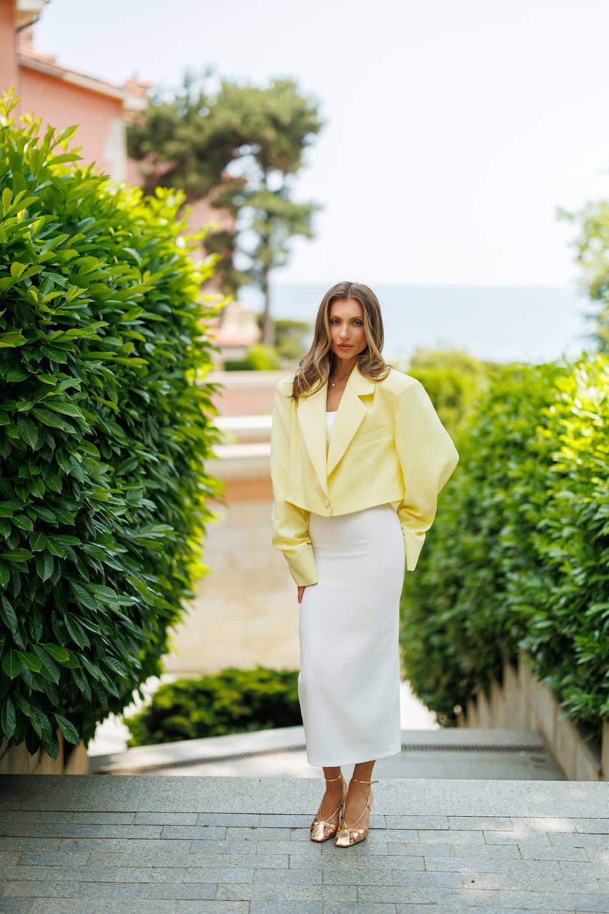 Woman in a yellow jacket and white skirt standing on a path with greenery and a clear sky.