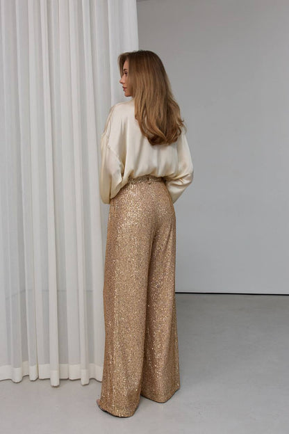 Woman wearing a white blouse and gold sequin pants standing in front of a white curtain.