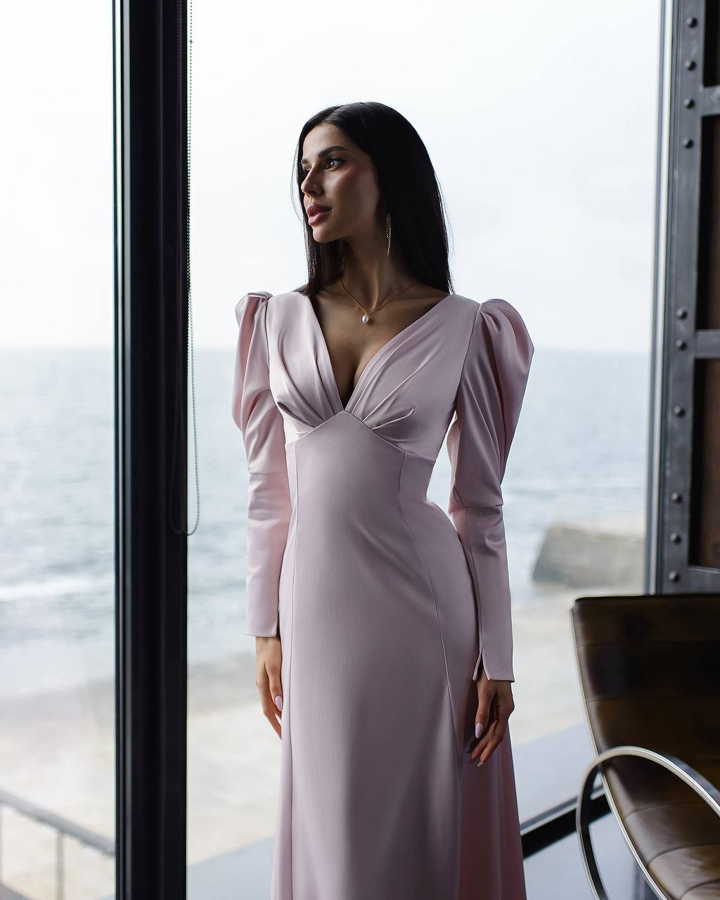 Woman in a pink dress standing by a large window with a view of the ocean.