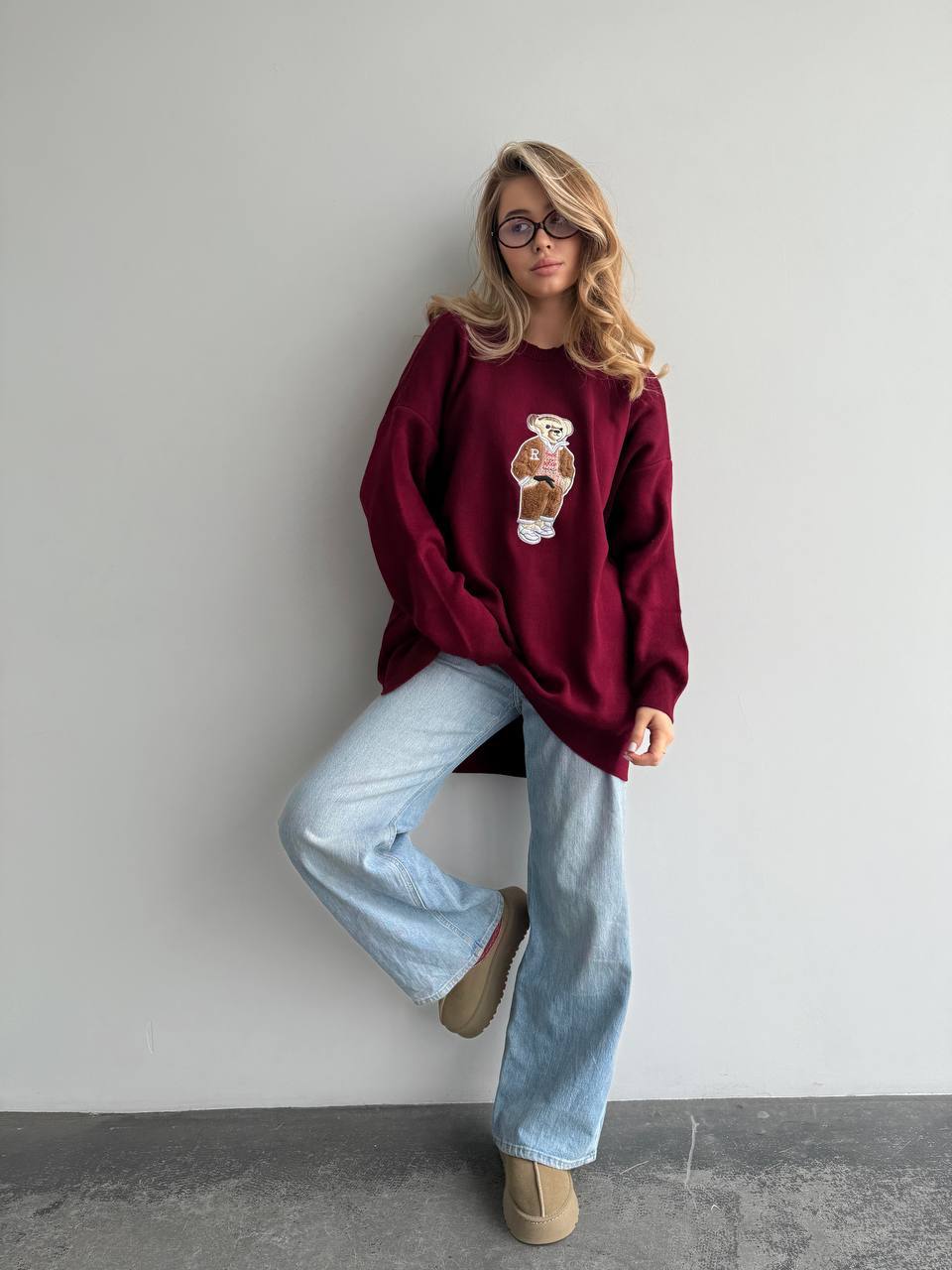 Person wearing a burgundy sweater with a graphic design, light blue jeans, and brown shoes against a plain background.