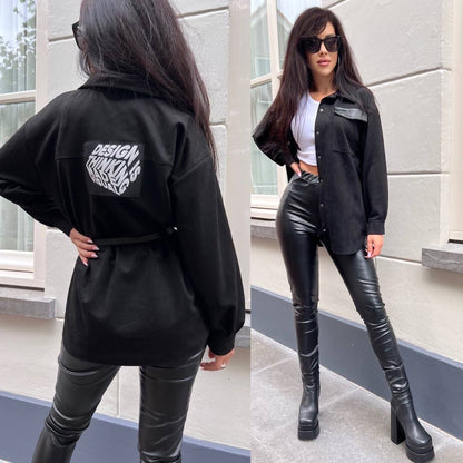 Woman wearing a black jacket with text on the back, white top, and black leather pants standing against a building.