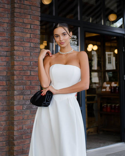 Woman in a white strapless dress holding a black clutch against a brick wall.